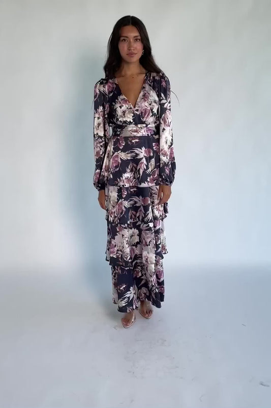 Model wearing the Montrouge Tiered Satin Maxi Dress in Navy + Lilac Floral featuring a V-neckline, short flutter sleeves, a smocked back, and a flowing tiered satin maxi skirt with a navy background and lilac floral print.