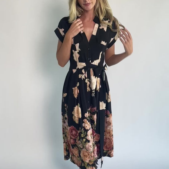 Model wearing a black floral midi dress with a collared neckline, functional button-front bodice, short sleeves, flap chest pockets, a removable waist tie, and a flowing midi-length skirt.