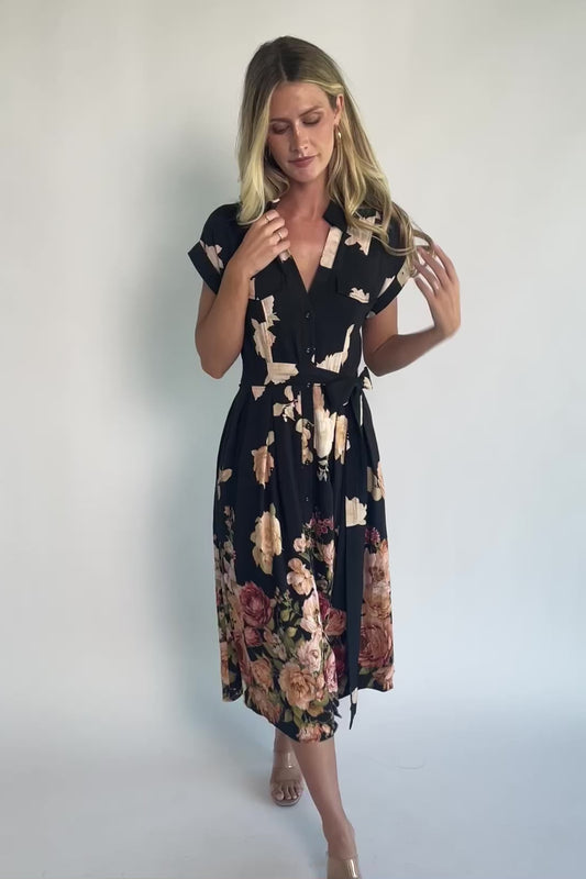Model wearing a black floral midi dress with a collared neckline, functional button-front bodice, short sleeves, flap chest pockets, a removable waist tie, and a flowing midi-length skirt.