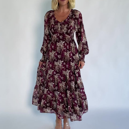 Model wearing a burgundy floral maxi dress with a V-neckline, smocked bodice, and long sheer sleeves with smocked ruffle cuffs.