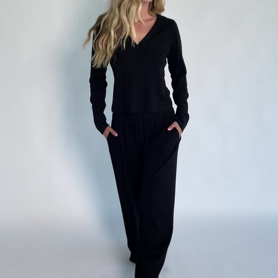 Model wearing the Rubi Soft Ribbed Pajama Set in Black featuring a soft ribbed knit, long-sleeve V-neck top with a snap-button placket, and matching straight-leg pants with an elastic waistband and side pockets.
