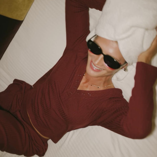 Model wearing the Rubi Soft Ribbed Pajama Set in Burgundy featuring a soft ribbed knit, long-sleeve V-neck top with a snap-button placket, and matching straight-leg pants with an elastic waistband and side pockets.