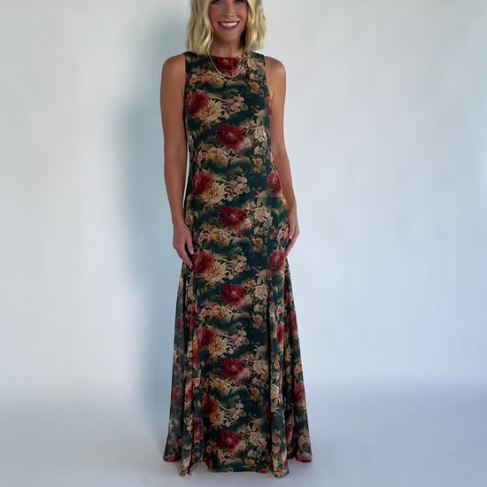 Model wearing a topaz floral maxi dress with a high round neckline, sleeveless bodice, and an open back with a sash tie.