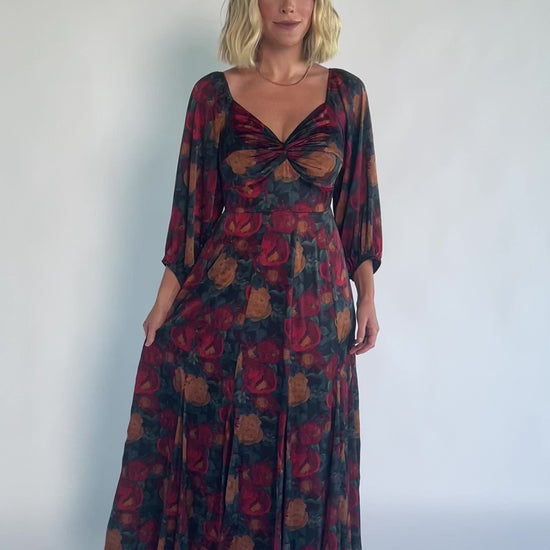 Model wearing a dark jade maxi dress with an oversized marsala floral print, a twist-knot V-neckline, half sleeves with elastic cuffs, a defined high waist, and a flowing maxi skirt with godet panels and an asymmetrical scalloped hem.