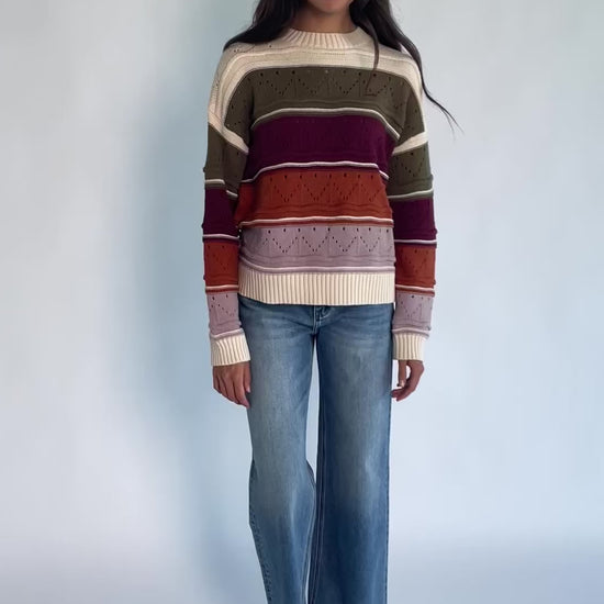 Ivory striped sweater with green, mulberry, rust, and purple horizontal stripes, a round neckline, dropped shoulders, long sleeves, and a relaxed fit.