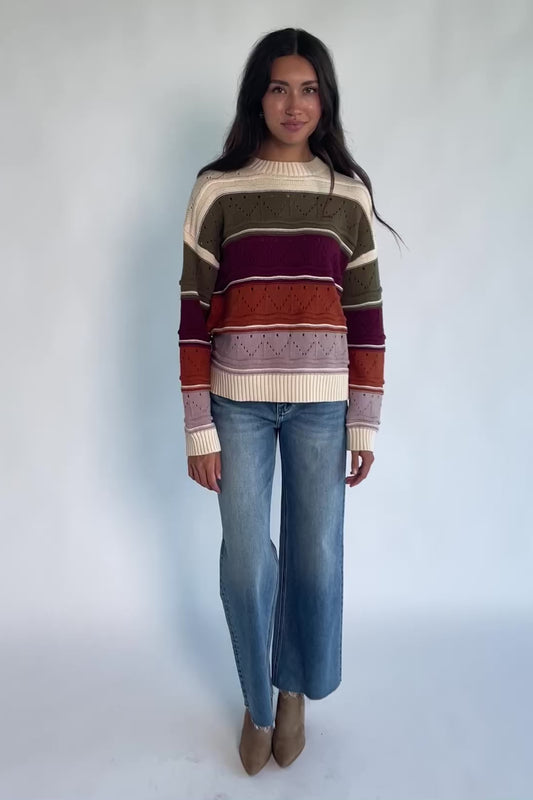 Ivory striped sweater with green, mulberry, rust, and purple horizontal stripes, a round neckline, dropped shoulders, long sleeves, and a relaxed fit.
