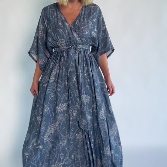 Model wearing a navy botanical-print maxi dress with a surplice V-neckline, kimono sleeves, an elastic waist with a removable sash tie, and a flowing full-length skirt featuring a side slit.
