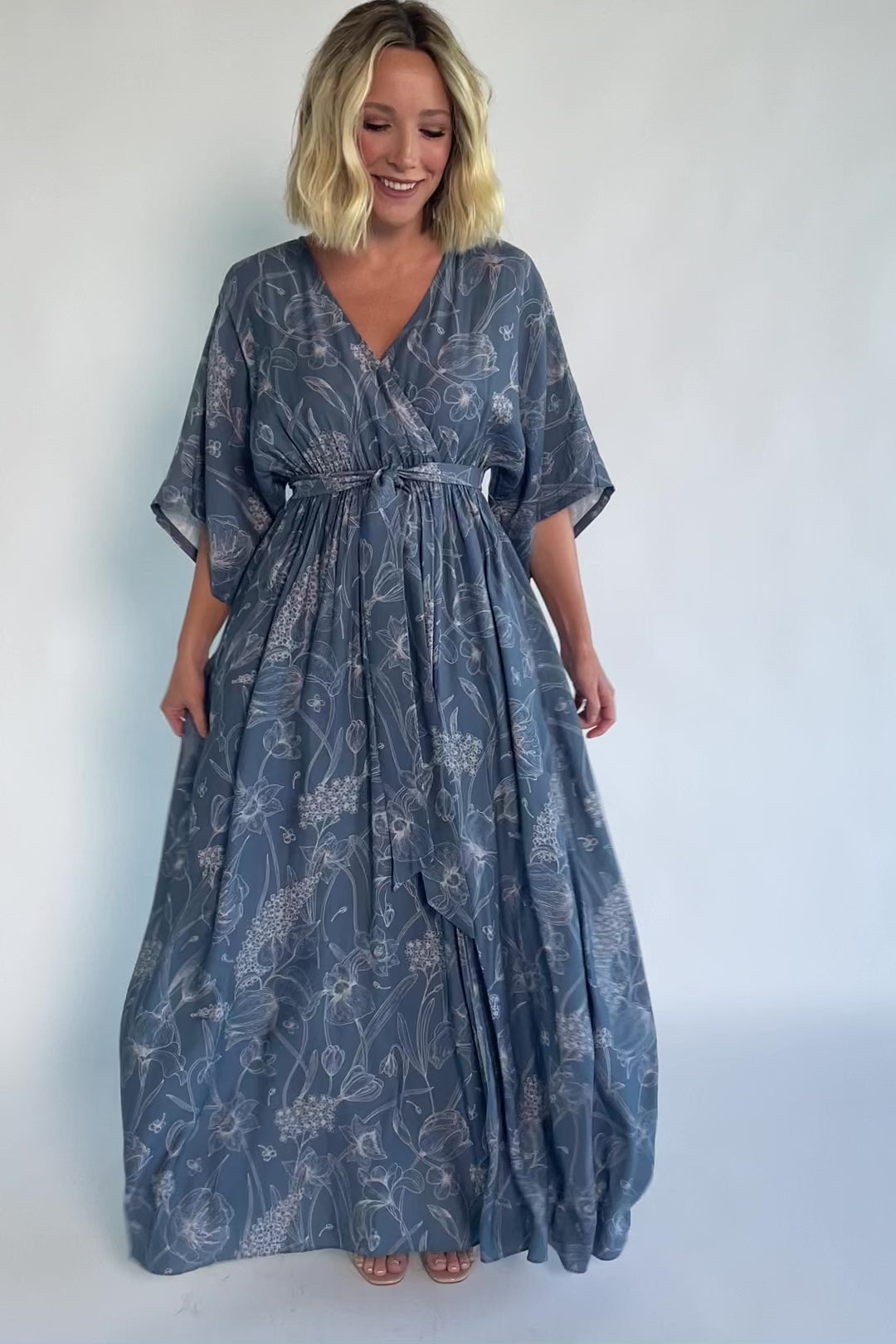 Model wearing a navy botanical-print maxi dress with a surplice V-neckline, kimono sleeves, an elastic waist with a removable sash tie, and a flowing full-length skirt featuring a side slit.