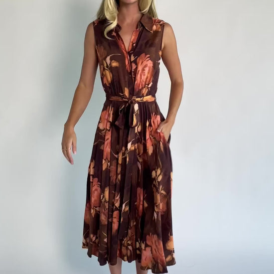Model wearing the Tess Midi Dress in Brown + Rust featuring a collared neckline, functional button-front bodice, sleeveless design, removable waist tie, elastic back waist, hidden side pockets, and a flowing midi skirt with a brown background and warm rust floral print.