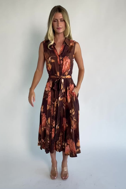 Model wearing the Tess Midi Dress in Brown + Rust featuring a collared neckline, functional button-front bodice, sleeveless design, removable waist tie, elastic back waist, hidden side pockets, and a flowing midi skirt with a brown background and warm rust floral print.