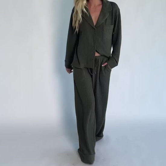 Model wearing a ribbed knit pajama set featuring a long-sleeve button-front V-neck top with a chest pocket and matching wide-leg pants with an elastic drawstring waist for a cozy, relaxed silhouette.