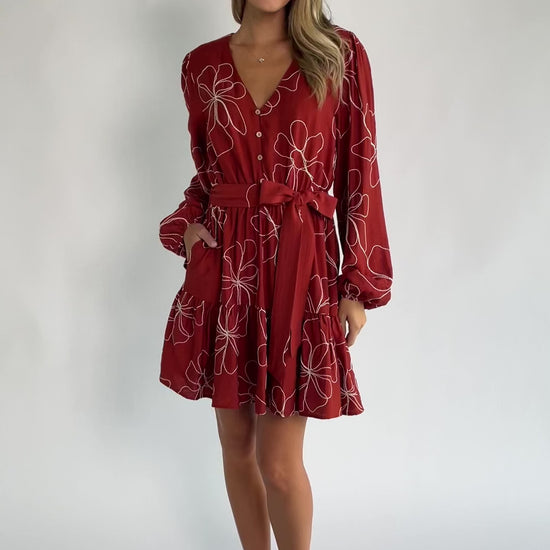 Rust floral mini dress with a V-neckline, button-front bodice, long balloon sleeves, elastic waistband with a self-tie sash, tiered ruffle hem, and side pockets.
