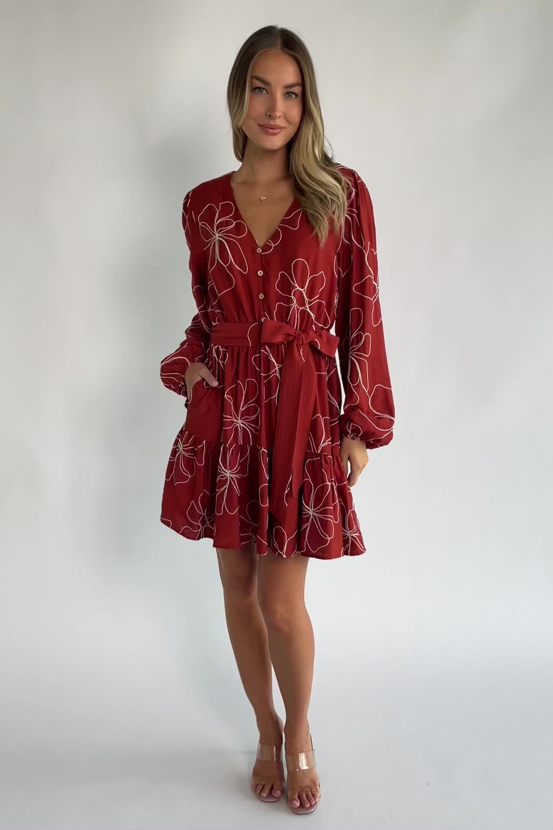 Rust floral mini dress with a V-neckline, button-front bodice, long balloon sleeves, elastic waistband with a self-tie sash, tiered ruffle hem, and side pockets.