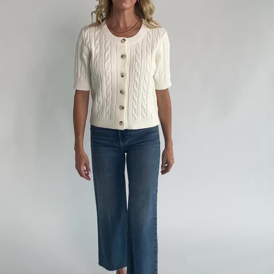 Cream cable knit cardigan with a round neckline, button-front closure, short sleeves, ribbed trim, and a fitted silhouette.