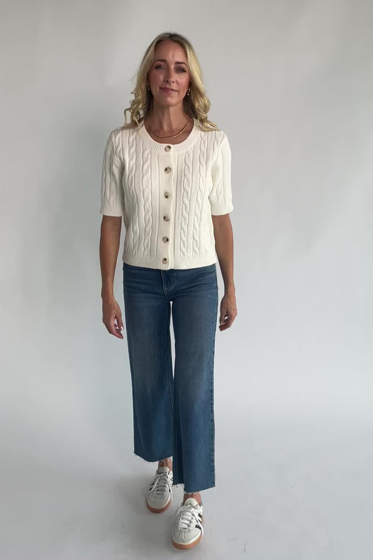 Cream cable knit cardigan with a round neckline, button-front closure, short sleeves, ribbed trim, and a fitted silhouette.
