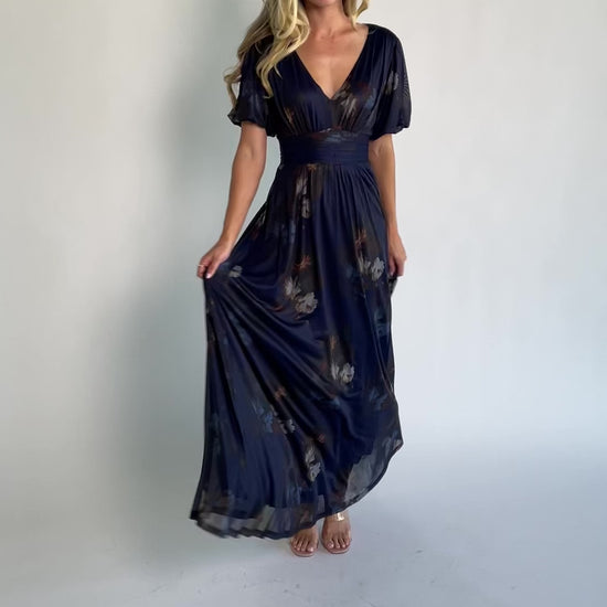 Model wearing the Whitney Mesh Maxi Dress in Navy, a navy floral mesh maxi dress with a deep V-neckline, short flutter sleeves, an empire waist, and a flowing A-line skirt with side pockets.