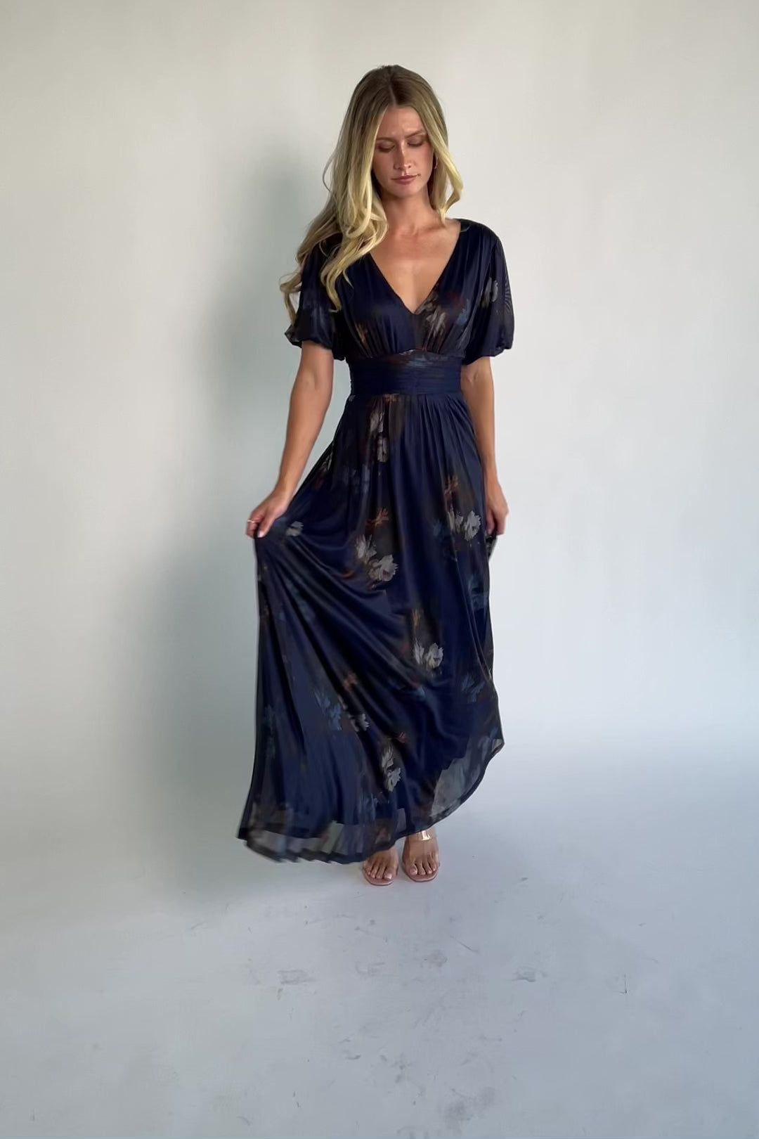 Model wearing the Whitney Mesh Maxi Dress in Navy, a navy floral mesh maxi dress with a deep V-neckline, short flutter sleeves, an empire waist, and a flowing A-line skirt with side pockets.