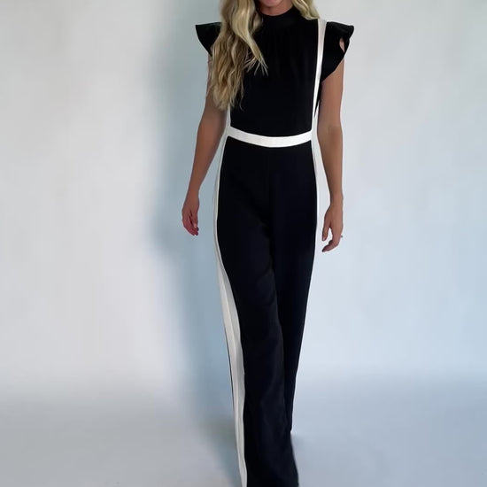 Model wearing the Lumina Ruffle Jumpsuit in Black + Ivory featuring a sleeveless ruffle-trim bodice, defined waist, and flowing wide-leg pants.
