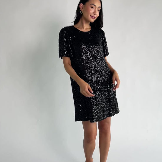 Black liquid sequin mini shift dress with a round neckline, short flutter sleeves, and a satin bow back detail.
