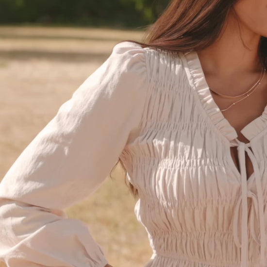 Marnie Smocked Top in Natural with smocked bodice, front bow ties, long sleeves, elastic cuffs, and peplum hem.
