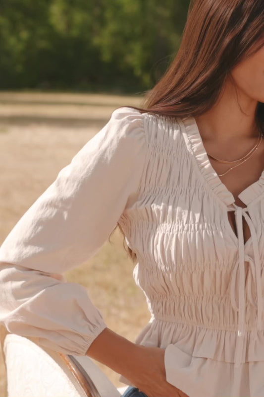 Marnie Smocked Top in Natural with smocked bodice, front bow ties, long sleeves, elastic cuffs, and peplum hem.