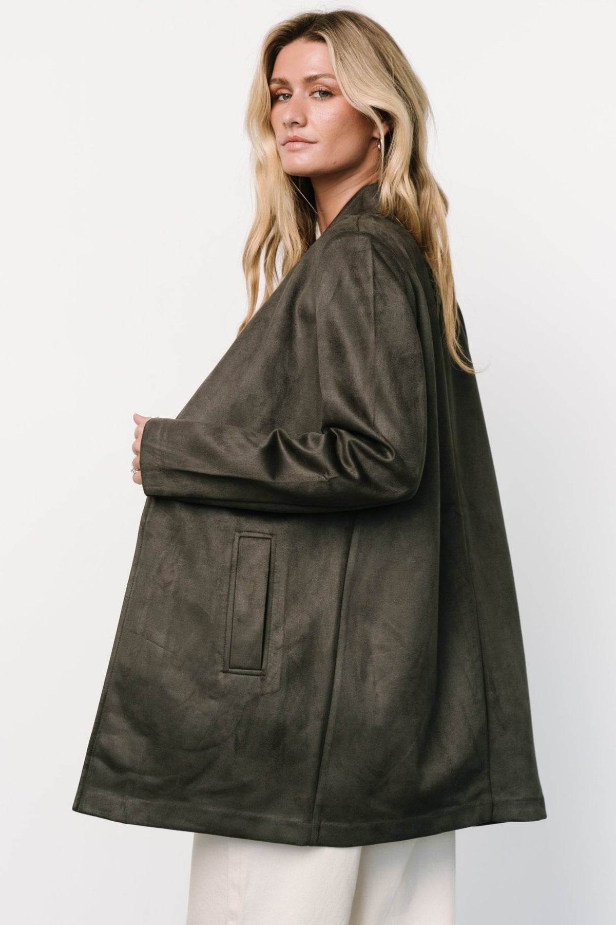 Model wearing charcoal faux-suede jacket showing side profile, thigh-length hem, long sleeves, and natural drape over outfit.