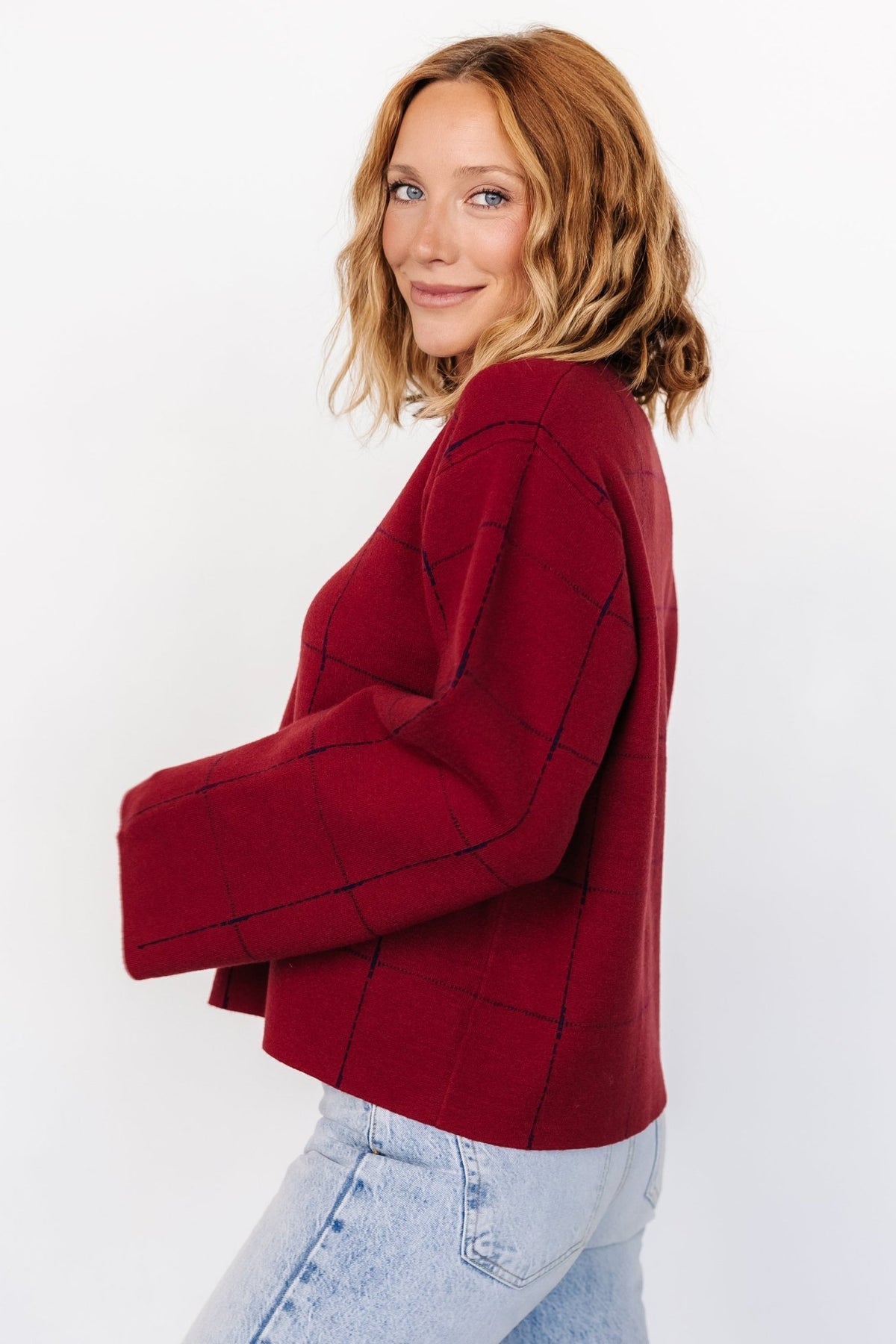 Three-quarter view showing the red sweater's navy windowpane detail and relaxed silhouette; medium-weight tight-knit texture visible on the body and sleeves.