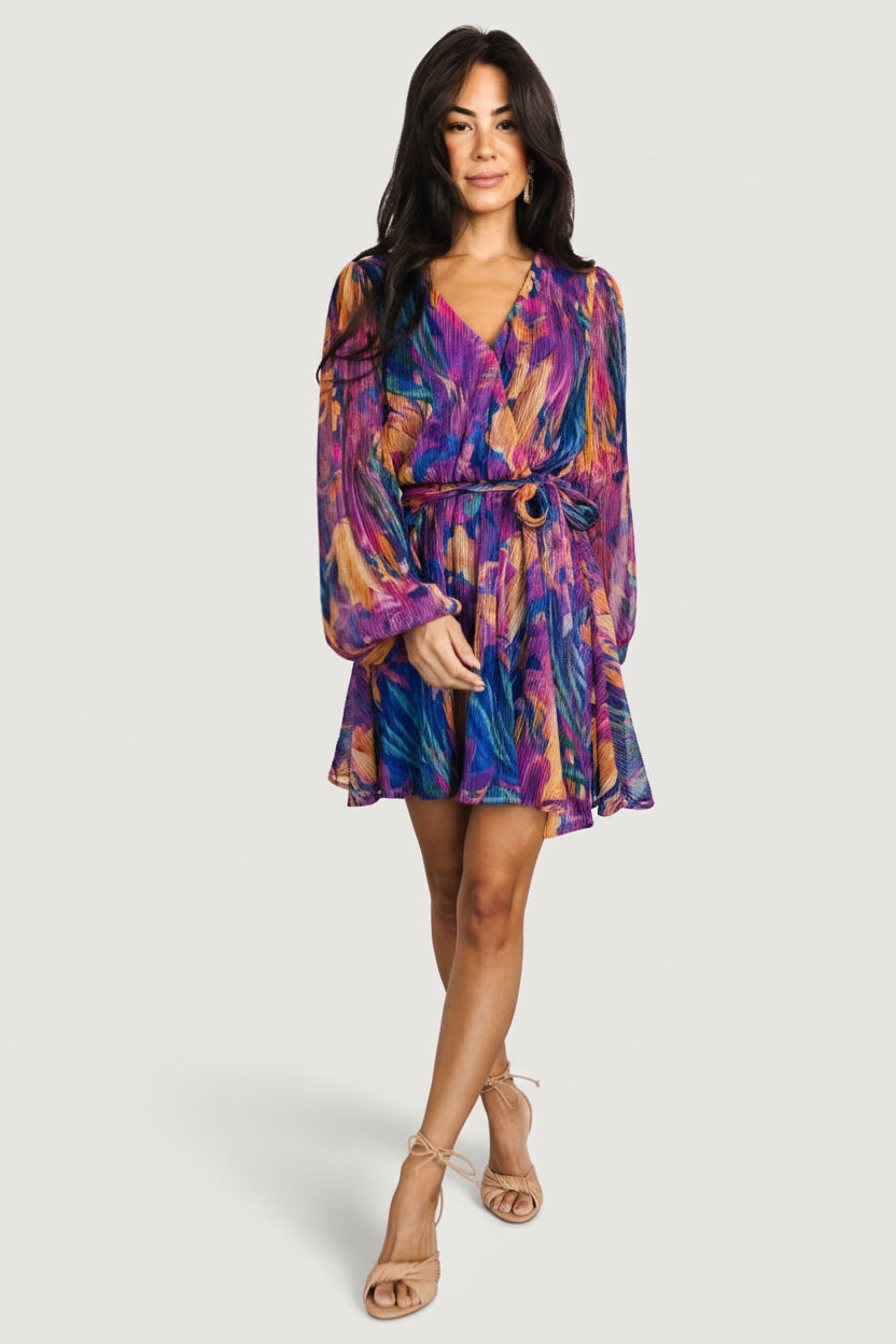 Front view of the Rev pleated mini dress in blue multi print on a model; crossover neckline, long sleeves, adjustable waist tie, mini hem.