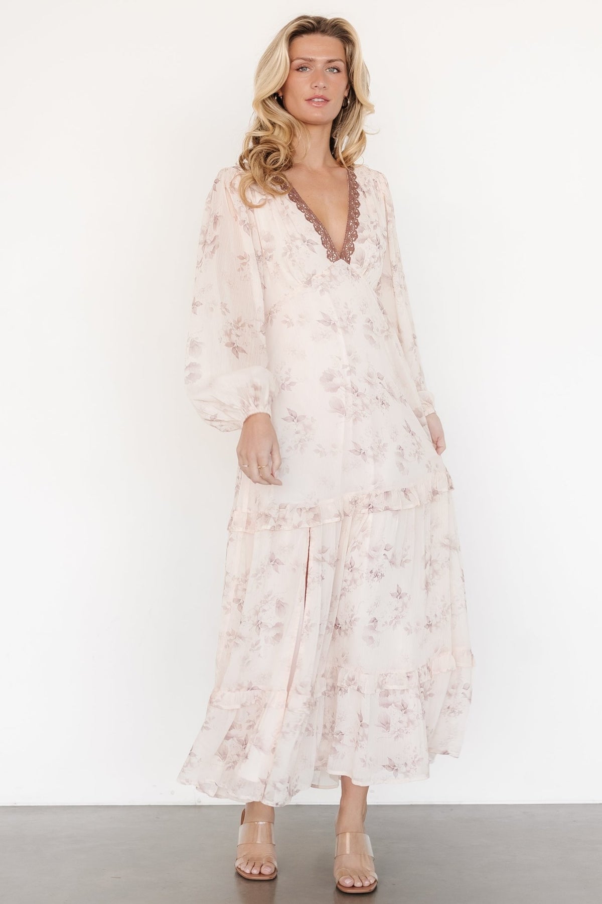 Three-quarter view highlighting the dress’s flowing two-tiered skirt, delicate beige and clove floral print, and sheer long sleeves with elastic cuffs.