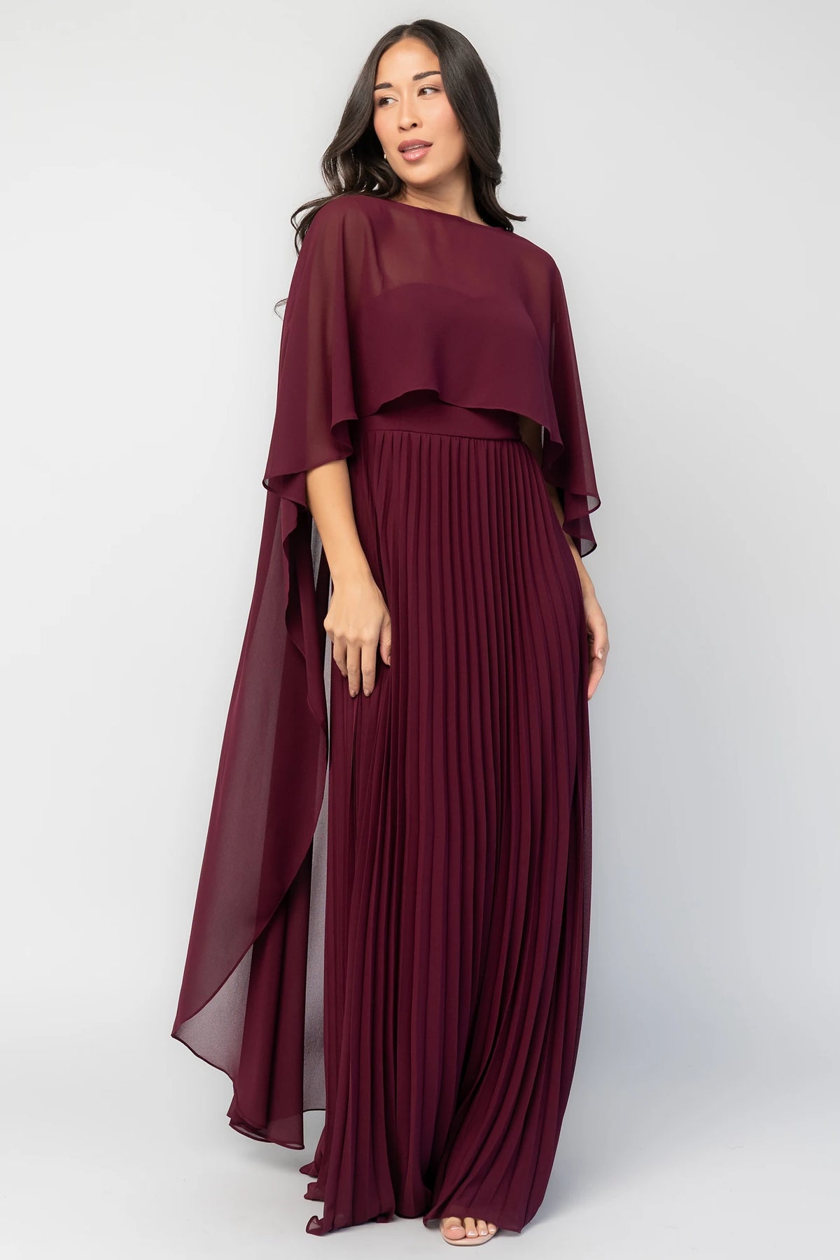 Serafina Strapless Gown in Burgundy with strapless neckline, removable sheer cape overlay, defined waist, and pleated skirt.