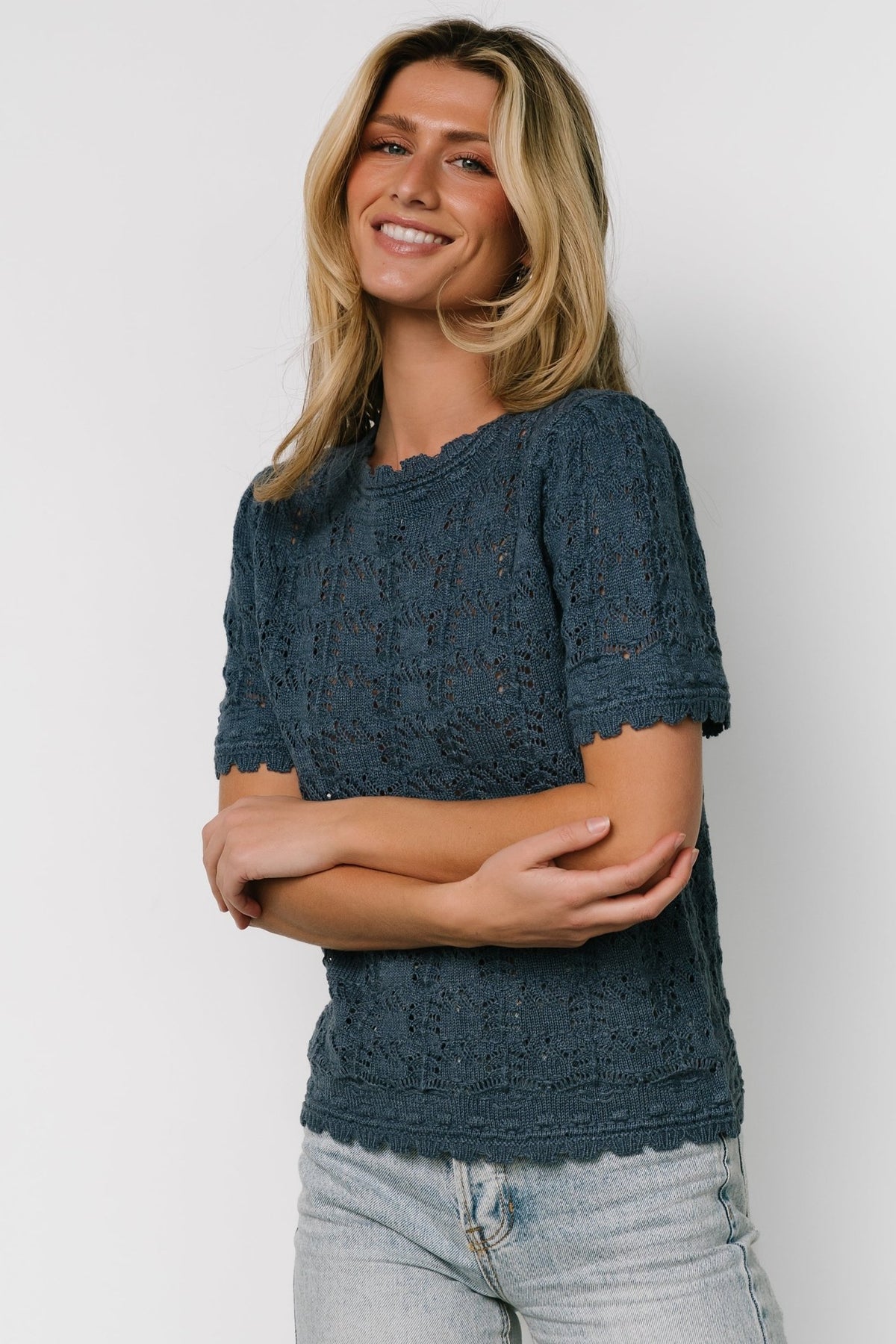 Side view of Serenity crochet knit sweater top in slate blue showing scalloped hem, fitted shoulder, and lightweight knit drape.