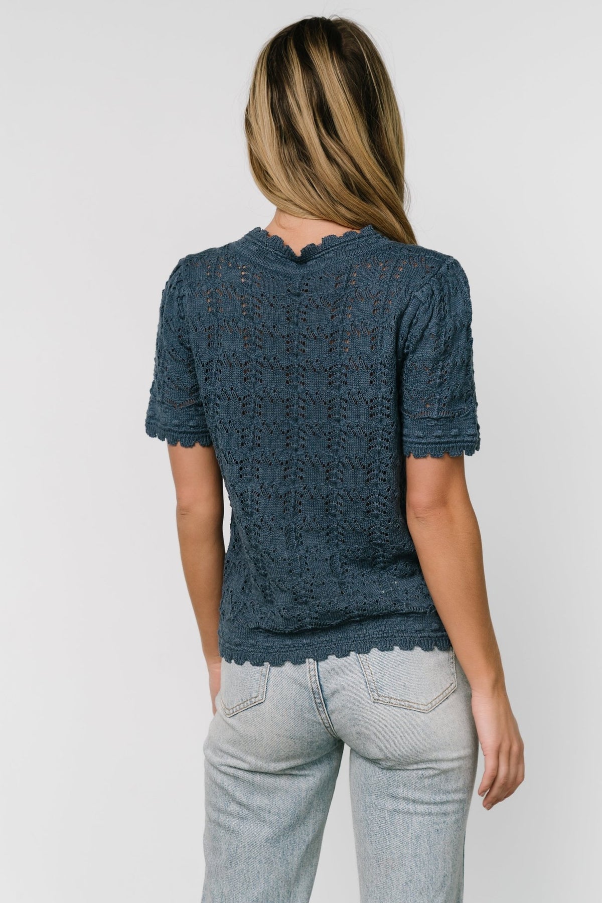 Back view of the Serenity crochet knit sweater top in slate blue displaying overall open-knit pattern and scalloped edge at hem.