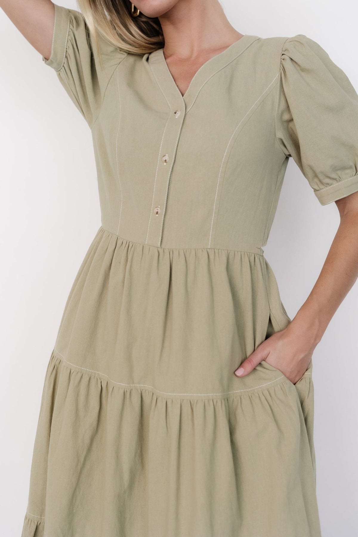 Model wearing sage green midi dress showing gathered shoulders, functional front buttons, white topstitching, and natural linen-like texture.