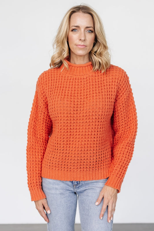 Front view of an apricot orange chunky knit sweater on a model, showing rolled funnel neck and wide-ribbed cuffs in soft waffle texture (product front).