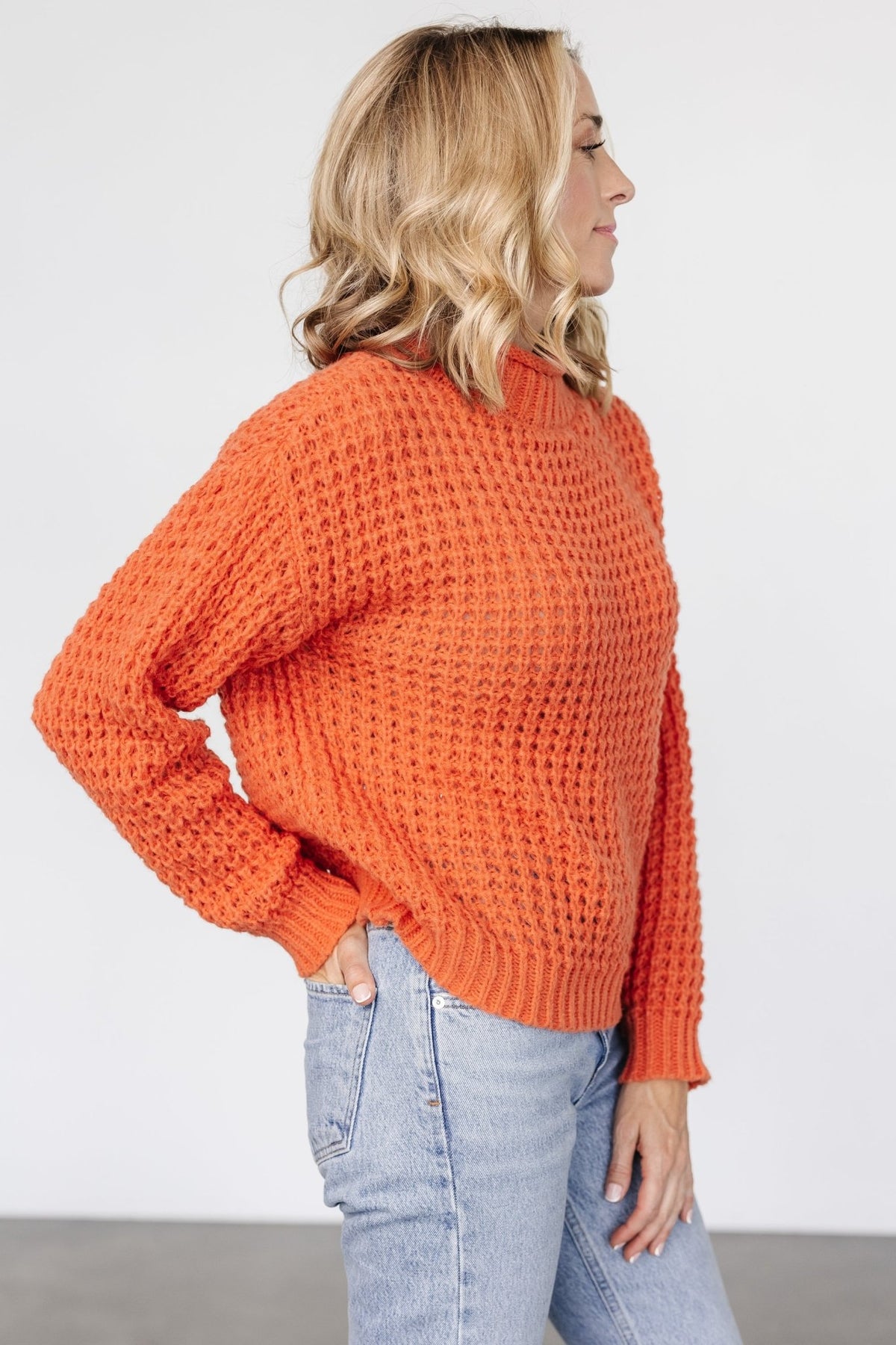 Side-angle shot of apricot orange sweater highlighting dropped shoulder and relaxed boxy silhouette with textured raised knit and wide hemline detail.