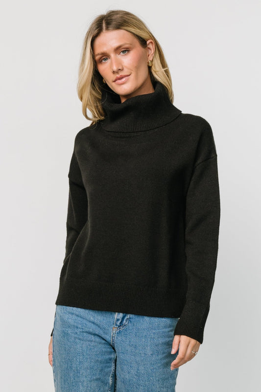 Front view of black turtleneck sweater on model, showing wide cowl neck, roomy silhouette, and ribbed cuff and hem details.