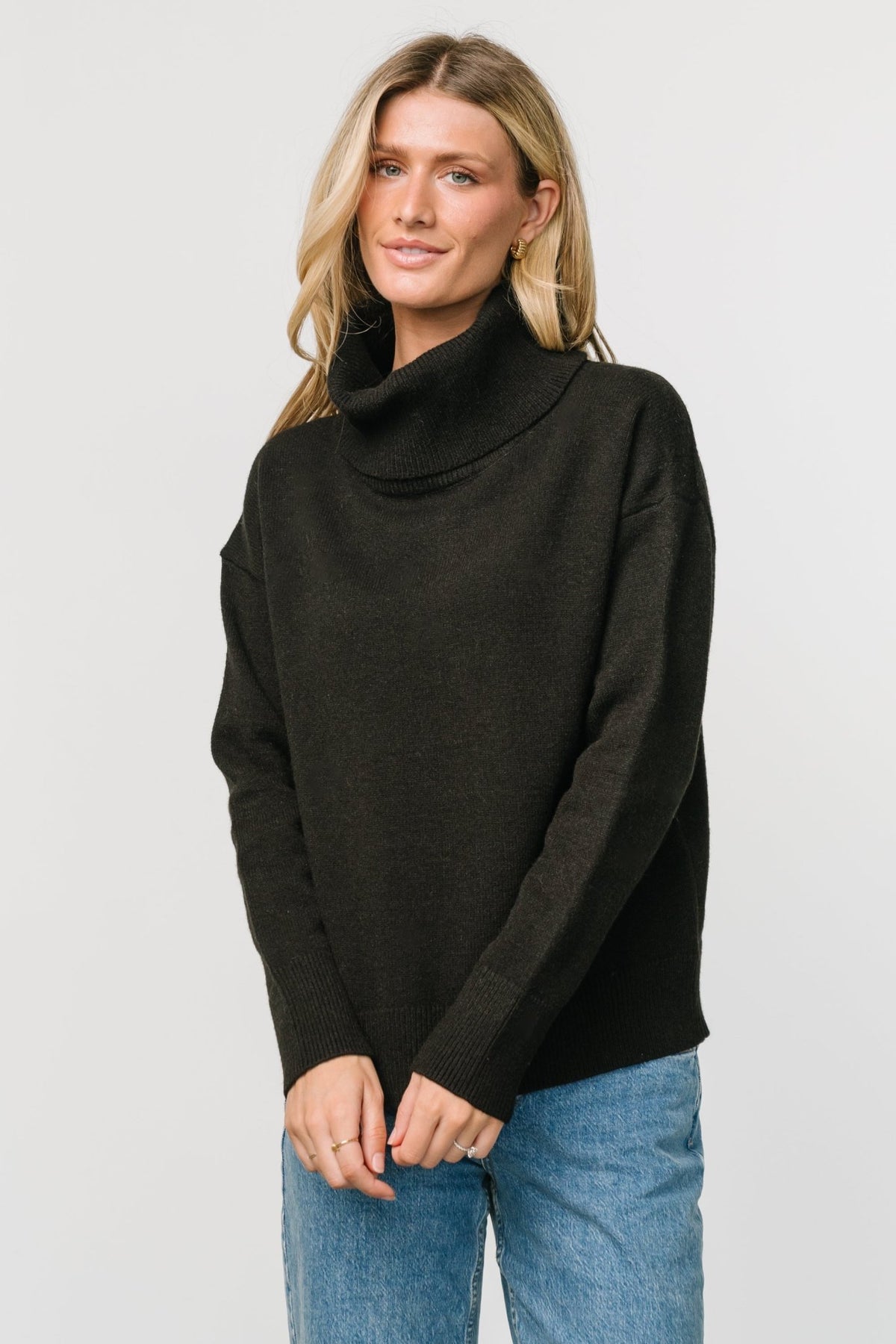 Model seated wearing black cowl turtleneck sweater styled with leggings to show drape and comfortable medium-weight knit.