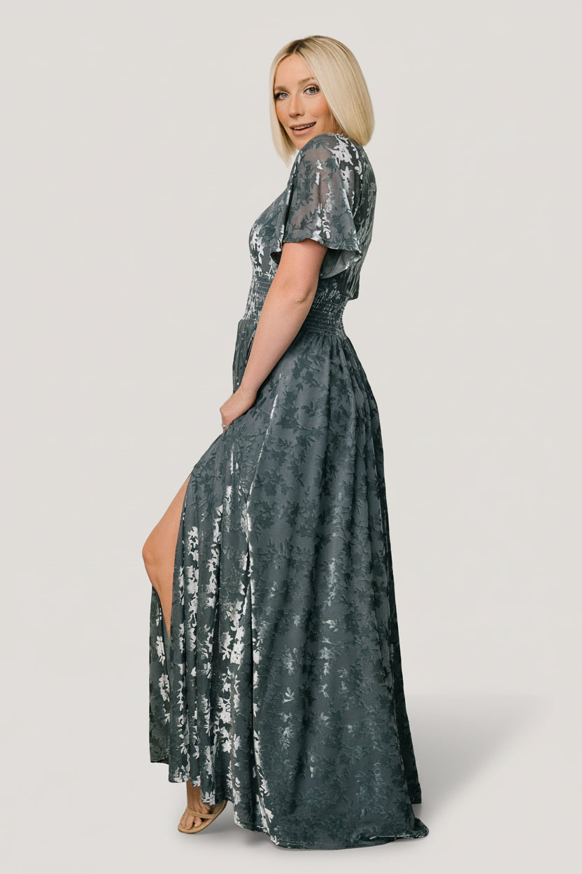 Model wearing Veronica velvet maxi dress in slate blue showing deep V-neckline and short flowy sleeves with the embossed velvet close-up.