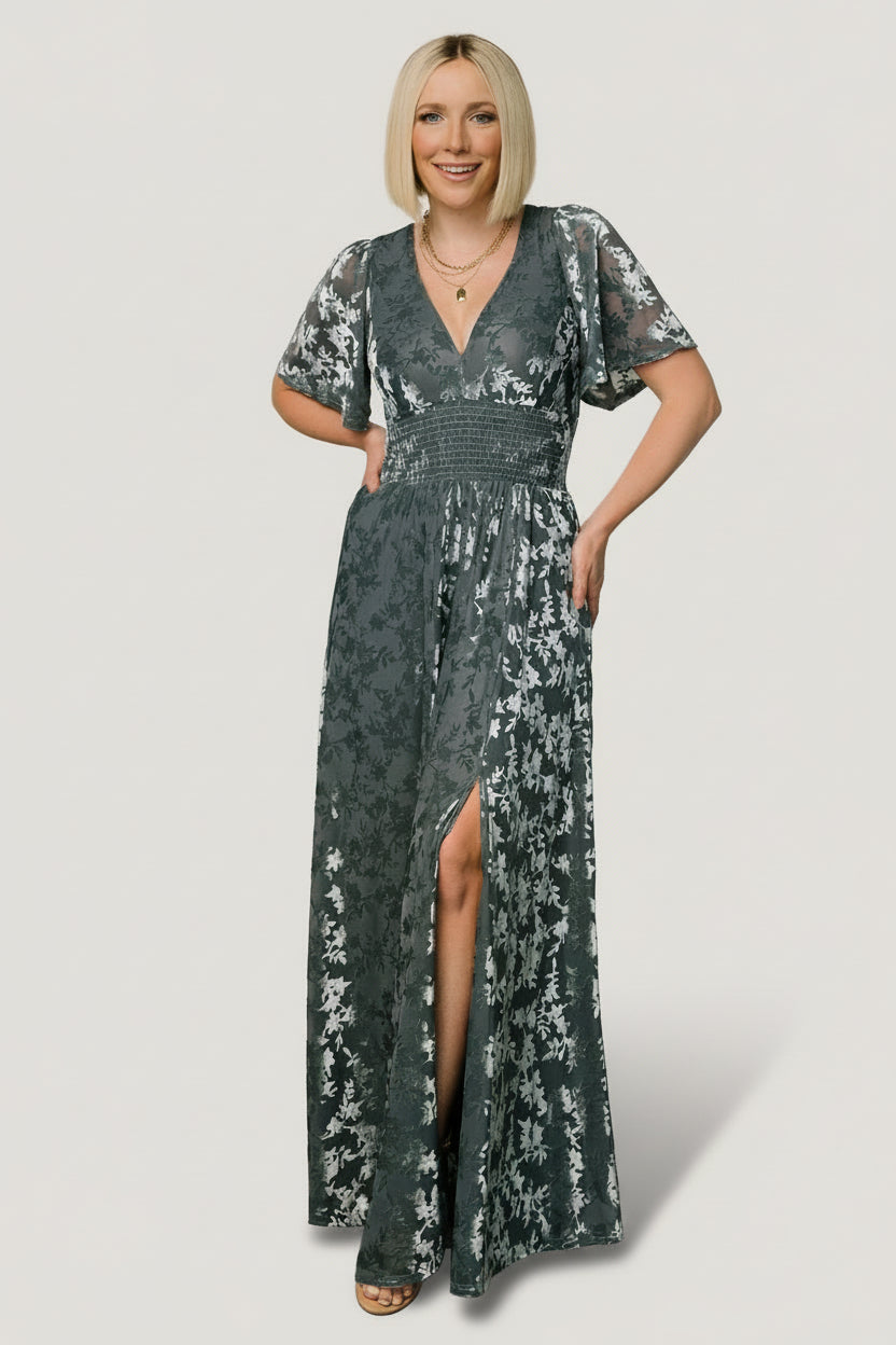 Front view of Veronica velvet maxi dress in slate blue showing deep V-neck, smocked waist, flowy sleeves, and left leg slit in soft embossed texture.
