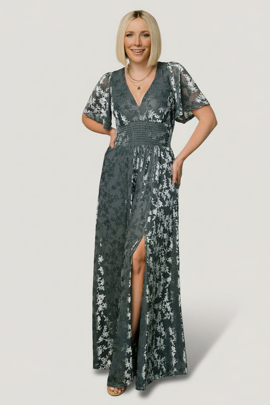 Front view of Veronica velvet maxi dress in slate blue showing deep V-neck, smocked waist, flowy sleeves, and left leg slit in soft embossed texture.