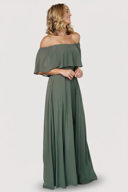 Model front view wearing dark sage off-shoulder maxi dress with elastic neckline, gathered waist and left-side slit, standing on plain background.