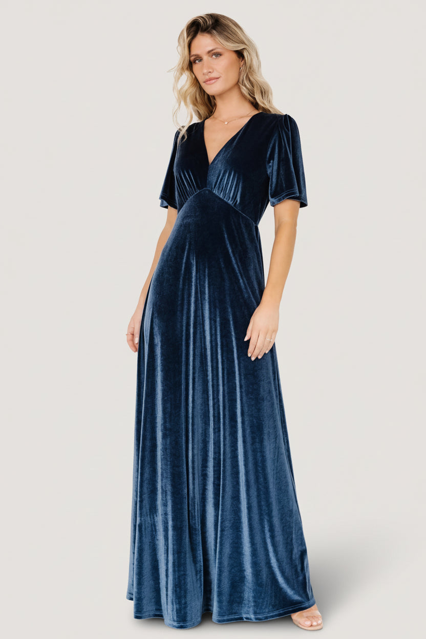 Side view of blue Vivian velvet maxi dress displaying flutter sleeve and empire waist seam, with fabric texture and subtle sheen visible in close framing.