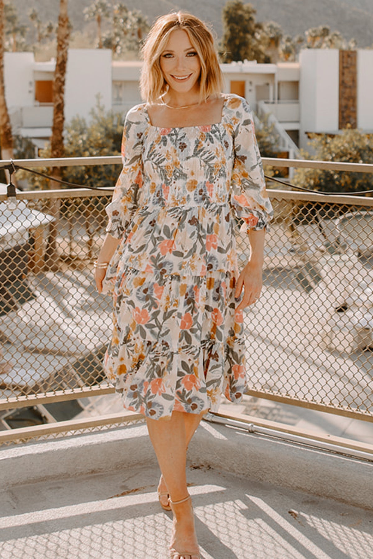 Model walking in a multi floral smocked midi dress demonstrating movement of the flowing tiered skirt and 3/4 ruffle sleeves.
