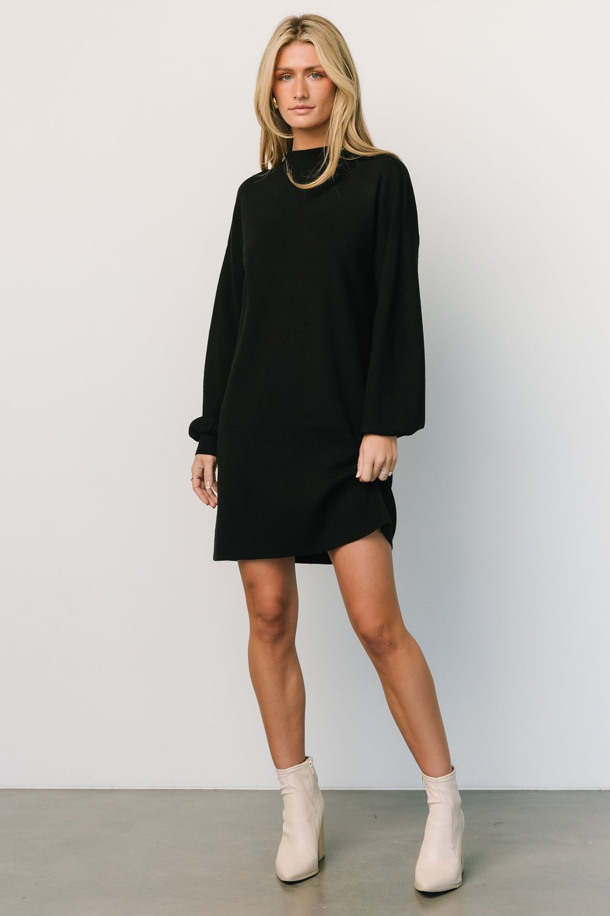 Front view of a short black sweater dress with mock neckline and long bubble sleeves on a model, styled with ankle boots and minimal accessories.