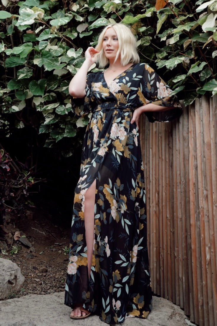 Model wearing the black floral maxi dress in a front pose displaying the elastic waist with a detached self-tie sash and side slit.