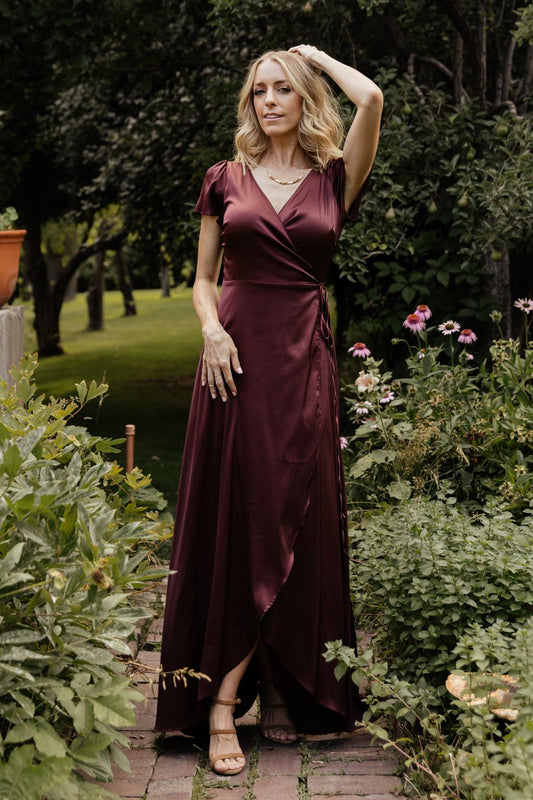 Model wearing Krystal Satin Wrap Gown in dark clove, front view showing V-neckline, tulip skirt and fluid satin drape across the body.