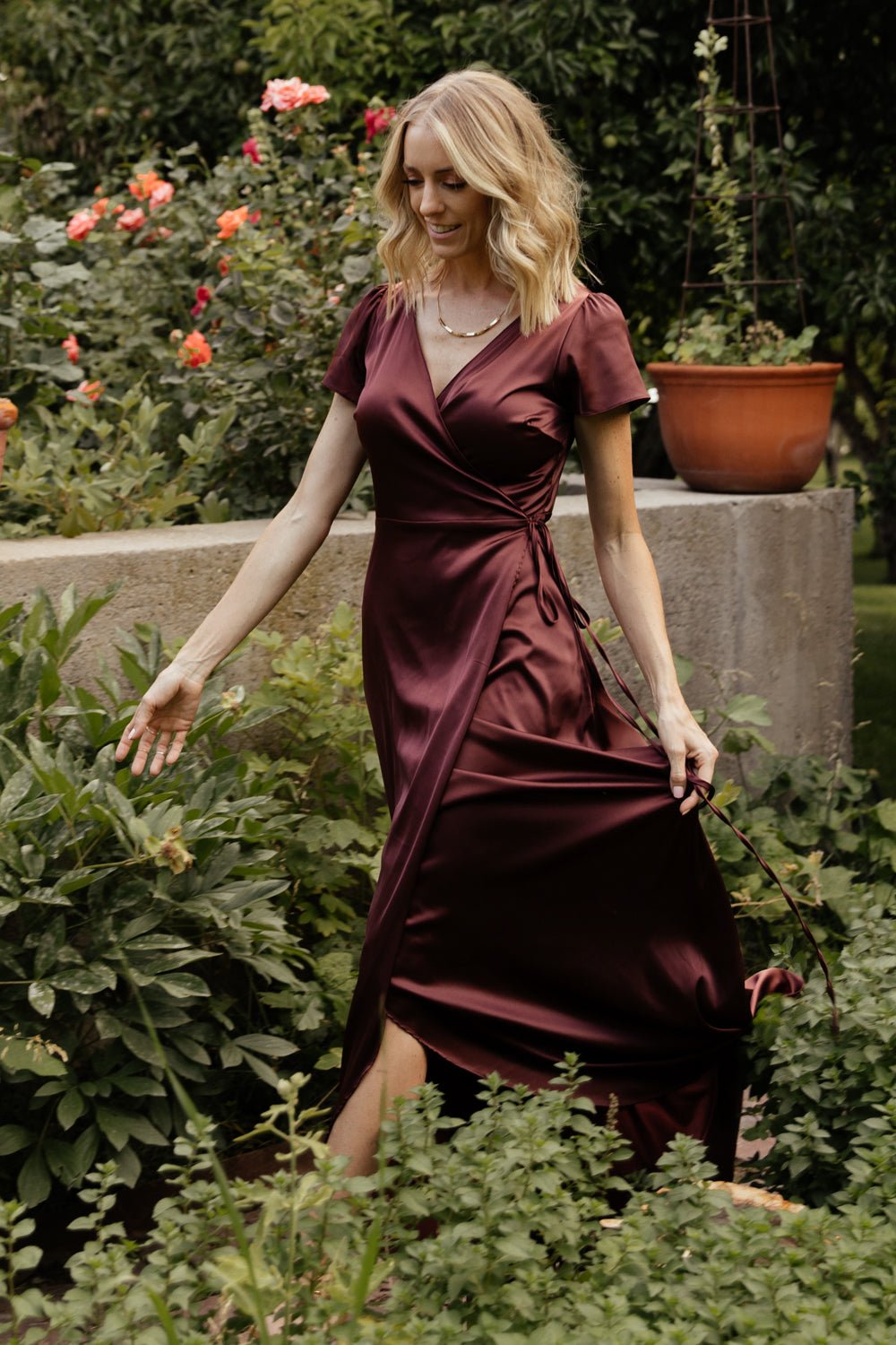 Three-quarter view of Krystal Satin Wrap Gown in dark clove highlighting flutter sleeve, side wrap tie, and gentle sheen of the fabric.