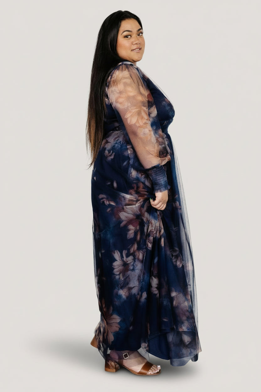 Model wearing dark blue floral tulle maxi dress, long sheer sleeves with smocked cuffs, and ankle-length hem detail.