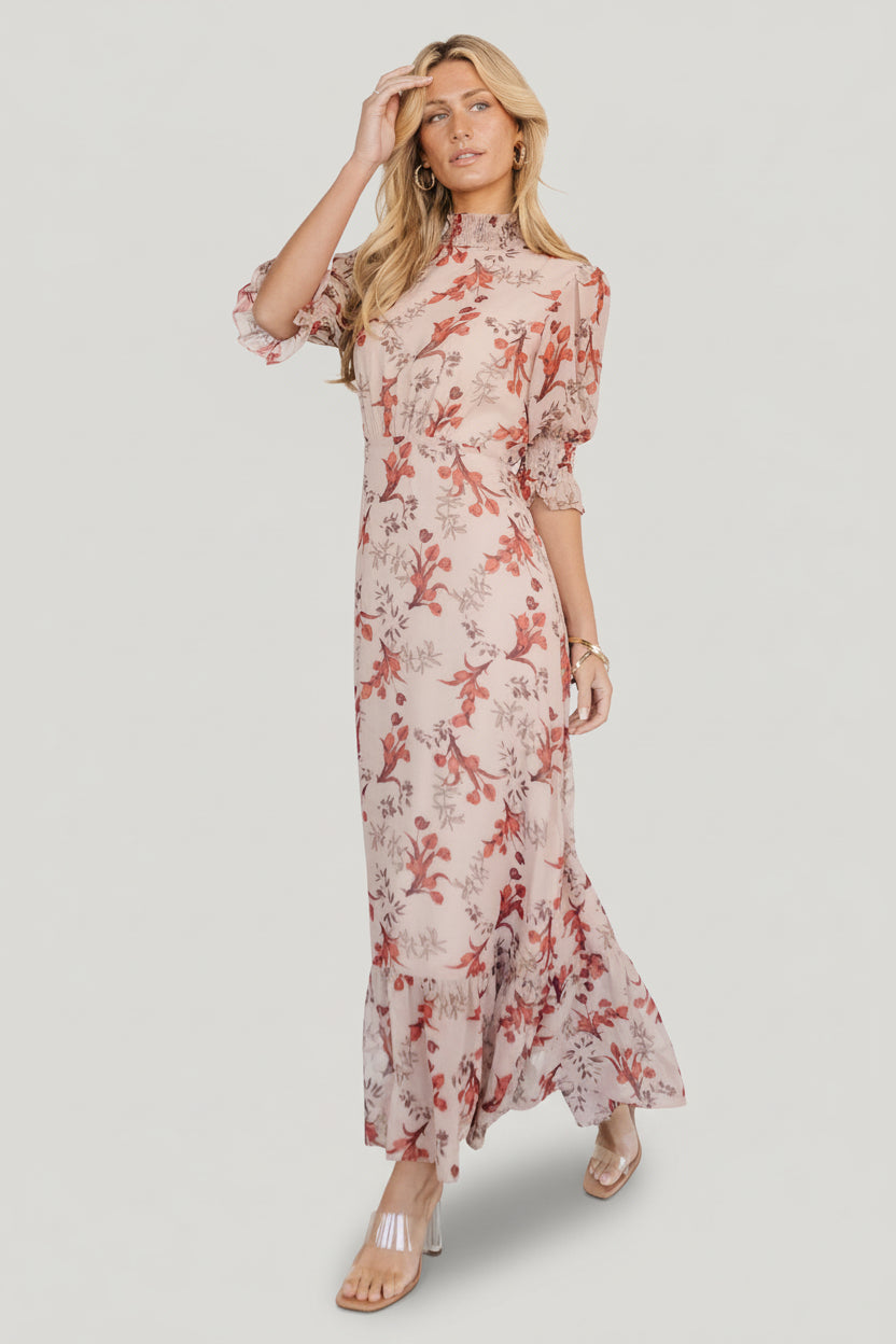 Front view of blush floral chiffon maxi dress on model, showing smocked mock neckline, pleated waist and single-tier flowing skirt in soft light.