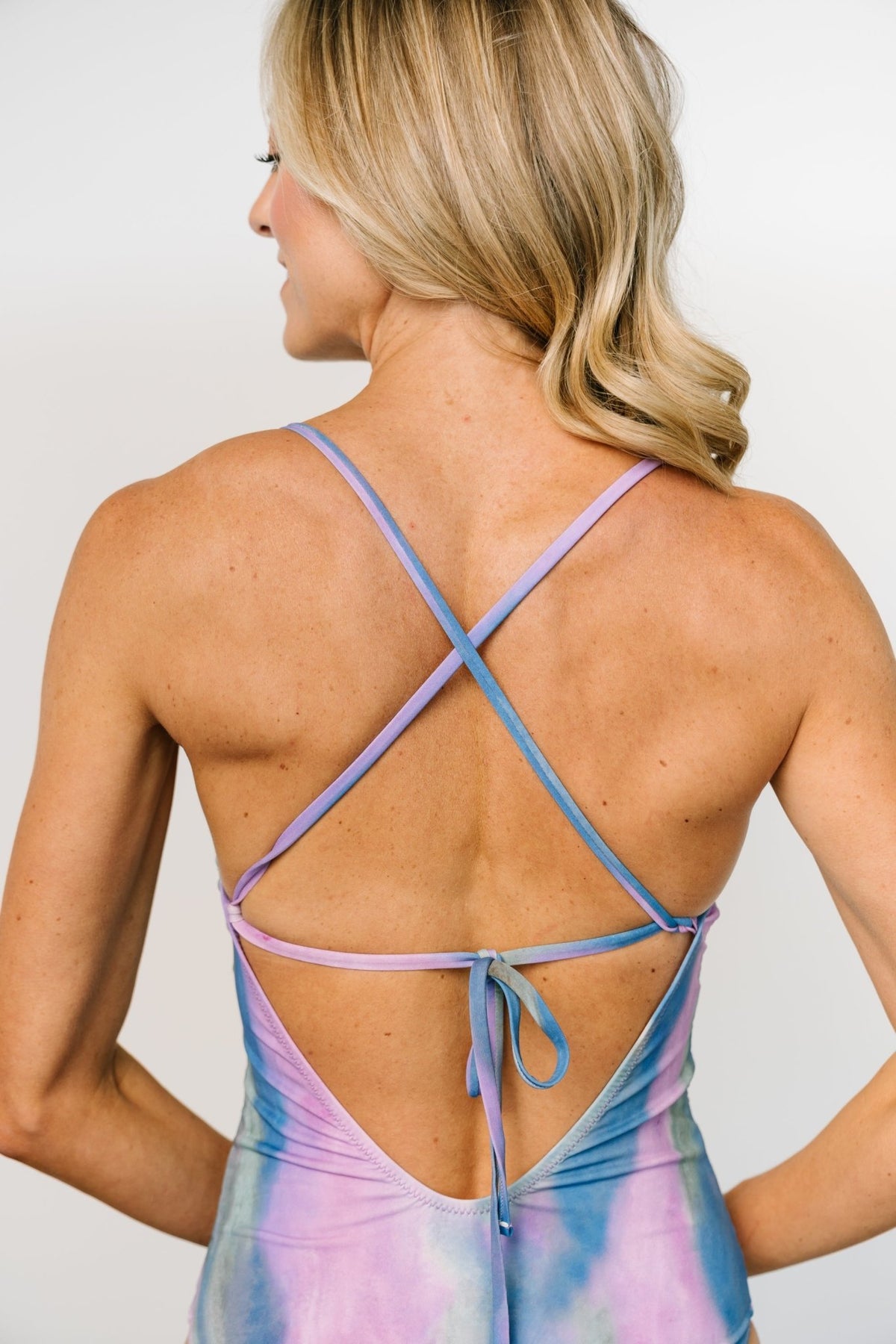 Detail shot of the adjustable tie and strap junction on the back of the purple-blue tie-dye one-piece, showing construction and finish.